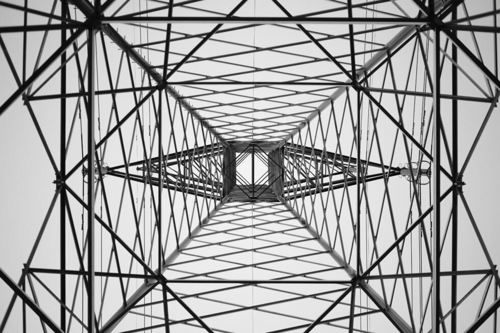 Alttan yukarı doğru çekilmiş, çelik bir elektrik iletim direğinin iç yapısını gösteren siyah-beyaz fotoğraf. Metal kirişler ve çapraz destekler merkezde geometrik, simetrik bir perspektif oluşturuyor.