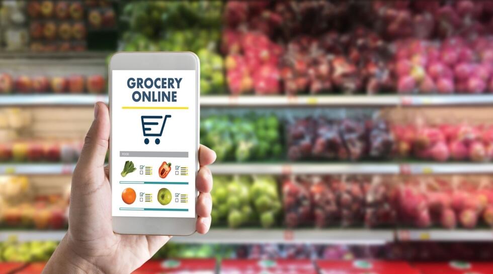 S&uuml;permarket reyonundaki taze meyve ve sebzelerin &ouml;n&uuml;nde, ekranda "Grocery Online" yazısı ve alışveriş sepeti simgesi bulunan akıllı telefonu tutan bir el.